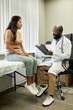 © TrueFrame Collective - Vertical full length shot of African American doctor wearing white coat listening to young adult Hispanic woman and writing down symptoms in health card