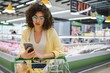 © Serhii - Young woman using smartphone while shopping in supermarket