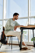 © TrueFrame Collective - Vertical image of young man sitting at desk and studying online using his laptop in the room with big windows