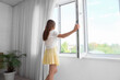 © Pixel-Shot - Young woman opening window in room, back view