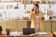 © LIGHTFIELD STUDIOS - Young woman stands in her cozy kitchen, casually talking on the phone and holding a drink.