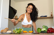 © sutulastock - Fit and smiling woman in a white sports top tasting fresh vegetable salad with chopsticks in a modern kitchen, surrounded by healthy food ingredients and vegetables.