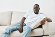 © SHOTPRIME STUDIO - A young African man in a casual white t shirt and jeans, relaxed on a couch, conveying confidence and comfort in a bright, minimalistic setting