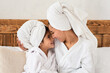 © Prostock-studio - Sweet little girl having beauty day with her mother at home or at spa hotel, wearing white bathrobes, wrapping towels all over their heads, sitting on bed and cuddling, enjoying time together