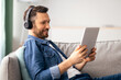 © Prostock-studio - Joyful middle-aged bearded man laying on couch in living room, usind wireless headset and digital tablet, listening to music or watching videos online, using mobile apps, copy space