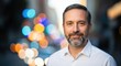 © Von - A man with a beard and mustache, wearing a white shirt, standing in front of a blurred cityscape with colorful lights.