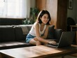 © onyengradar - Young Asian woman working on laptop at home while sitting on sofa in cozy living room