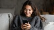 © Johannes - happy millennial hispanic teen girl checking social media holding smartphone at home smiling young latin woman using mobile phone app playing game shopping online ordering delivery relax on sofa no l