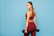 © ViDi Studio - Back side view young fitness trainer instructor sporty woman sportsman wear red sportswear training in home gym hold in hand dumbbells isolated on plain blue background. Workout sport fit abs concept.