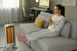 © New Africa - Young woman with cup of drink sitting on sofa near electric heater at home