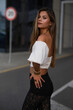 © Евгений Бордовский - Stylish young woman in elegant black lace pants and a white off-shoulder top posing outdoors, leaning on a railing with an urban background in warm evening light.
