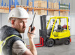 © Grispb - Warehouse worker in safety helmet and vest uses walkie talkie near forklift truck, logistics, cargo handling, transportation. Logistics, Warehouse, Transportation.