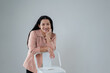 © pichet - Smiling woman leaning on a chair in a modern and minimalistic environment, dressed in a stylish light pink blouse with a friendly expression and long black hair