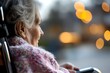© Yuliia - Senior Caucasian woman in wheelchair wearing cozy knit scarf gazes thoughtfully through window with soft golden bokeh lights creating peaceful, contemplative atmosphere.