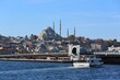 A scenic view of the Süleymaniye Mosque rising above Istanbul’s skyline, with the Galata Bridge and a ferryboat crossing the Golden Horn under a bright blue sky.