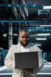 © DC Studio - Engineer in server farm managing infrastructure systems, checking recovery plan on laptop. Data center worker monitoring performance using automation tools, making sure rigs are functioning optimally