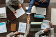 © pressmaster - Top view showing group of middle aged multiethnic business professionals collaborating at table, two people shaking hands over laptop and financial charts, others reviewing documents