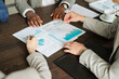 © pressmaster - Caucasian woman and Black man discussing financial charts and graphs during business meeting, hands gesturing over printed documents on wooden table, top view composition
