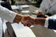 © pressmaster - Caucasian woman and Middle aged Black man shaking hands over desk, both wearing business attire, documents and digital tablet visible on table, top view emphasizing professional agreement
