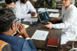 © pressmaster - Middle aged Caucasian man sitting at table with diverse group of doctors discussing medical documents and laptop during meeting, hands clasped under chin, top view