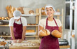 © JackF - Professional young female baker presenting fresh baked loaves of bread with satisfied smile, standing in warm inviting bakery filled with variety of baked goods