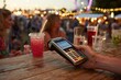 © whitestorm - Close-up of hand holding payment terminal at a lively outdoor festival bar.