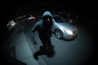 © alexkoral - male burglar thief in a hood breaks car in a parking at night. Video recording from a street surveillance system. Car robbery. CCTV