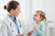 © AnnaStills - Caucasian female doctor smiling while interacting with Caucasian toddler girl in medical office, doctor holding medical documents, child looking up at doctor with cheerful expression