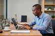 © AnnaStills - Young adult Black man working on laptop at desk in modern office, focusing on typing with documents and coffee cup nearby, demonstrating professional business environment