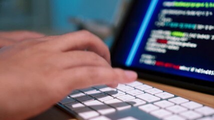 A close-up of a person's hands typing on a white keyboard, with a blurred computer screen in the background displaying lines of colorful code.