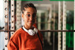 © Studio Marmellata - A young Black woman with braided hair and glasses smiles confidently at the camera. She wears an orange shirt and white headphones in a modern gym environment.