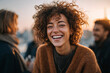 © Nano Photos - Joyful woman with curly hair laughing outdoors at sunset with friends gathered in the blurry background.