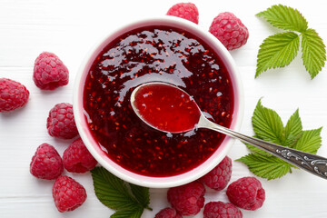 Wall Mural - Sweet raspberry jam in bowl, berries and green leaves on white wooden table, flat lay