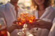 © Arcner - Close-up of two women and one man toasting with Aperol Spritz in sunshine, wearing white shirts, sharp focus on hands, shallow depth of field, warm tone