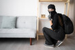 © Andrii Lysenko - Female Burglar or thief in black balaclava stealing money from apartment showing shh silence gesture. Criminal, house crime and breaking. robbery and burglary on property.
