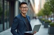 © Vadym - Young Asian businessman with laptop outdoors in modern city. Confident, smiling man in suit with glasses represents success, technology, connectivity. Urban pro lifestyle, entrepreneurship, career