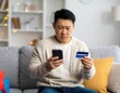 © Syafiq Rahman - Concerned man on sofa using phone, reviewing credit card, possibly facing online payment issues.