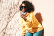 © halayalex - Beautiful woman with afro curls hairstyle.Smiling model in yellow hoodie. Sexy carefree female posing on the street background in sunglasses. Looking at smartphone screen, using apps
