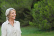 © wavebreak3 - Senior female standing smiling in grassy field by evergreen trees wearing linen blouse, copy space