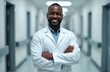 © Pete - Smiling black male doctor in professional white clothes stands looking at camera arms folded on chest, blurred hospital corridor background. Confident african american physician at work. Telemedicine