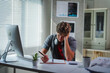 © PaeGAG - Exhausted male student working on laptop, writing notes near coffee mug in home workspace with computer and desk
