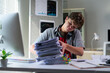 © PaeGAG - Young professional drowning in paperwork, looking stressed and overwhelmed at cluttered workspace, experiencing work-related anxiety