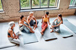 © HBS - Diverse women are sitting on mats in the studio, relaxing and chatting after a workout. Friends at the fitness studio.