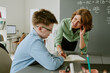 © AnnaStills - Caucasian middle aged woman leaning over desk assisting Caucasian teenage boy with schoolwork in classroom setting, both focused on open notebook, chalkboard with math equations in background