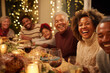 © Vitali - A diverse group of family members laughing heartily around a brightly lit dining table during a festive holiday meal.