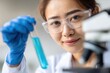 © oksa_studio - Scientist wearing lab coat and gloves analyzing blue liquid in test tube in laboratory next to microscope