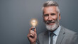 © buraratn - Smiling older man with beard holds glowing light bulb, symbolizing ideas and innovation. He wears suit and stands against gray background, exuding confidence and creativity