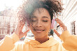 © halayalex - Beautiful woman with afro curls hairstyle.Smiling model in yellow hoodie.Sexy carefree female enjoying listening music in wireless headphones.Posing on street background at sunset