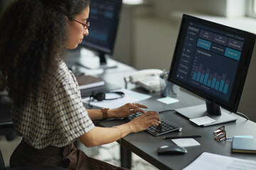 Wall Mural - Young adult woman working at desk using desktop computer with data analytics dashboard on screen, typing on keyboard, sitting in modern office environment, focused on task