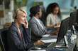 © DragonImages - Caucasian middle aged woman wearing headset working at computer in modern office, sitting at desk with two colleagues in background using computers and headsets